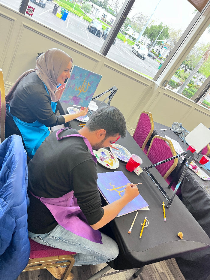 Two people sitting at a table with art supplies, including paint and brushes, in a room with large windows.