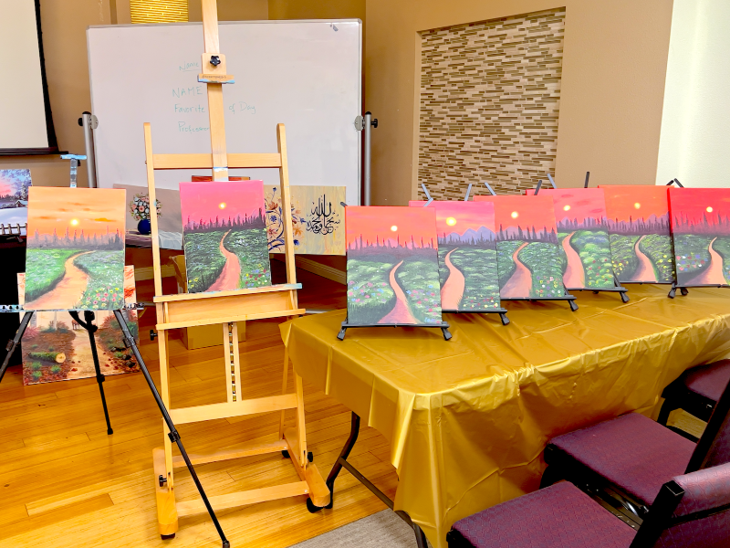 Paintings on easels and tables in a room with chairs and a screen.